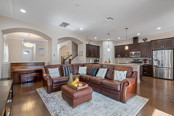 A spacious living room with a brown leather sofa, a wooden piano, and a fruit tray on an ottoman. - Beautiful Home Located in Best Location of Tampa (Tampa)
