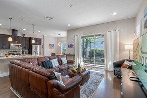 A modern living room with a brown leather sectional sofa, a dining area with dark wood cabinets and a built-in microwave, and a large sliding glass door leading to a balcony.