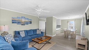 A living room with a blue sofa, a coffee table, and a ceiling fan. There is a dining area with a table and chairs.