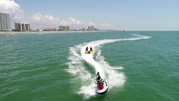 Un jet ski remolca una balsa amarilla con dos personas, otro jet ski con una persona y un horizonte urbano al fondo.