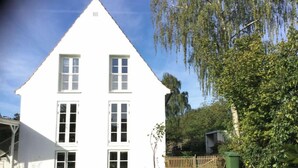 A white two-story house with a triangular roof, a front porch, and a small garden area with a lawn and a few plants.