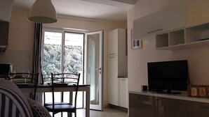 Een moderne keuken met een eethoek, een televisie en uitzicht op bomen door de open deur.