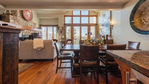 A spacious dining area with a wooden table and chairs, a fireplace, and large windows offering a view of the outdoors. - Classic Aspen Ridge Home by InvitedHome | Walk to Ski and Core, Wilson Views (Mountain Village)