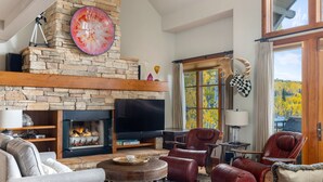 A cozy living room with a stone fireplace, leather chairs, a flat-screen TV, and a view of autumn foliage through large windows. - Classic Aspen Ridge Home by InvitedHome | Walk to Ski and Core, Wilson Views (Mountain Village)