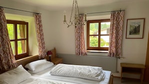 A bedroom with two beds, a nightstand, a lamp, a window with curtains, a radiator, and a picture on the wall. - Cozy vacation apartment with a unique view of the mountains (Ruhpolding)