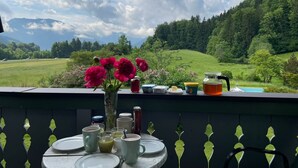 A table with a vase of flowers, various food items, and a glass of juice on a balcony overlooking a green landscape with mountains in the distance. - Cozy vacation apartment with a unique view of the mountains (Ruhpolding)