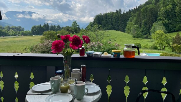 A table with a vase of flowers, various food items, and a glass of juice on a balcony overlooking a green landscape with mountains in the distance. - Cozy vacation apartment with a unique view of the mountains (Ruhpolding)