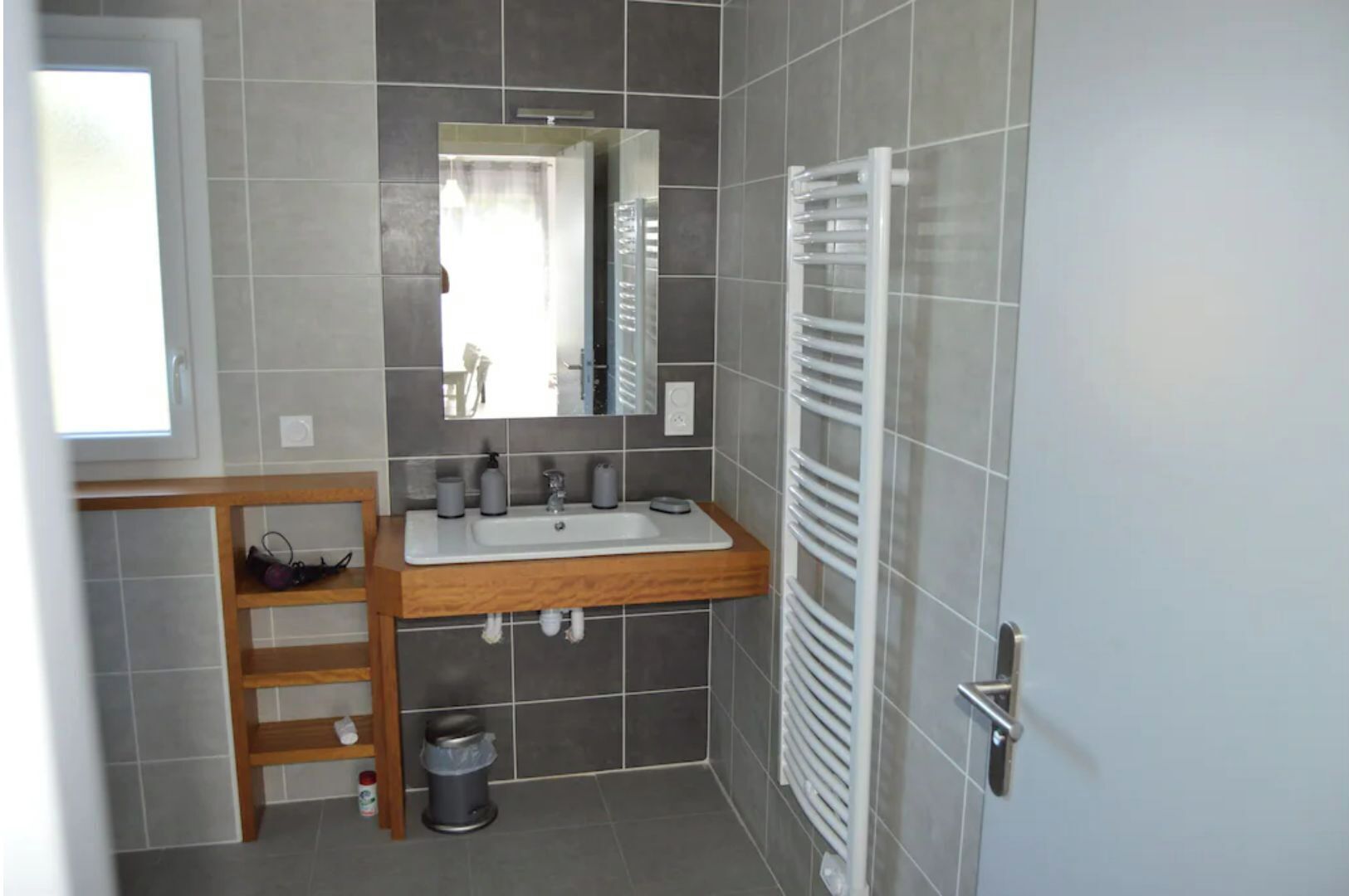 A bathroom with a white sink, mirror, and towel rack. There is a wooden shelf with toiletries and a trash can.