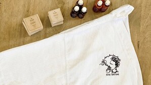 Um roupão branco com um logotipo, um quadro emoldurado e três frascos de produtos de banho sobre uma superfície de madeira.