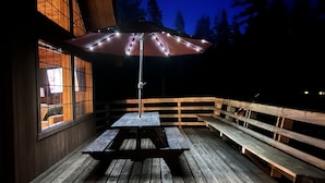 An outdoor wooden deck with a picnic table and benches, illuminated by string lights. - Little Bear Lodge Ponderosa Ca (Springville)