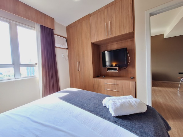 Habitación de hotel con cama, televisor montado en un mueble de madera y ventana con cortinas.