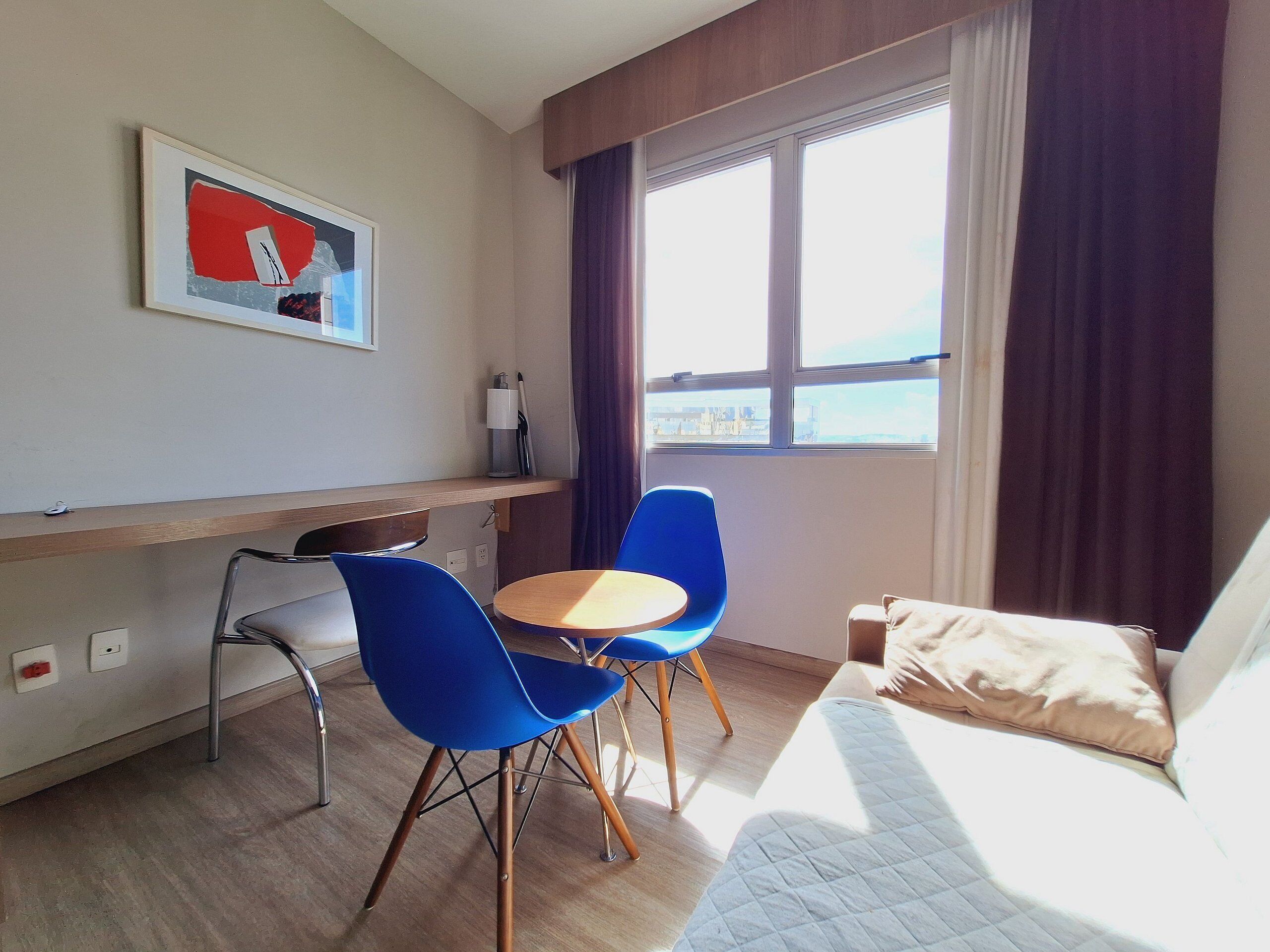 Habitación de hotel con una cama, un escritorio con una silla, una mesita, una ventana con cortinas y un cuadro en la pared.