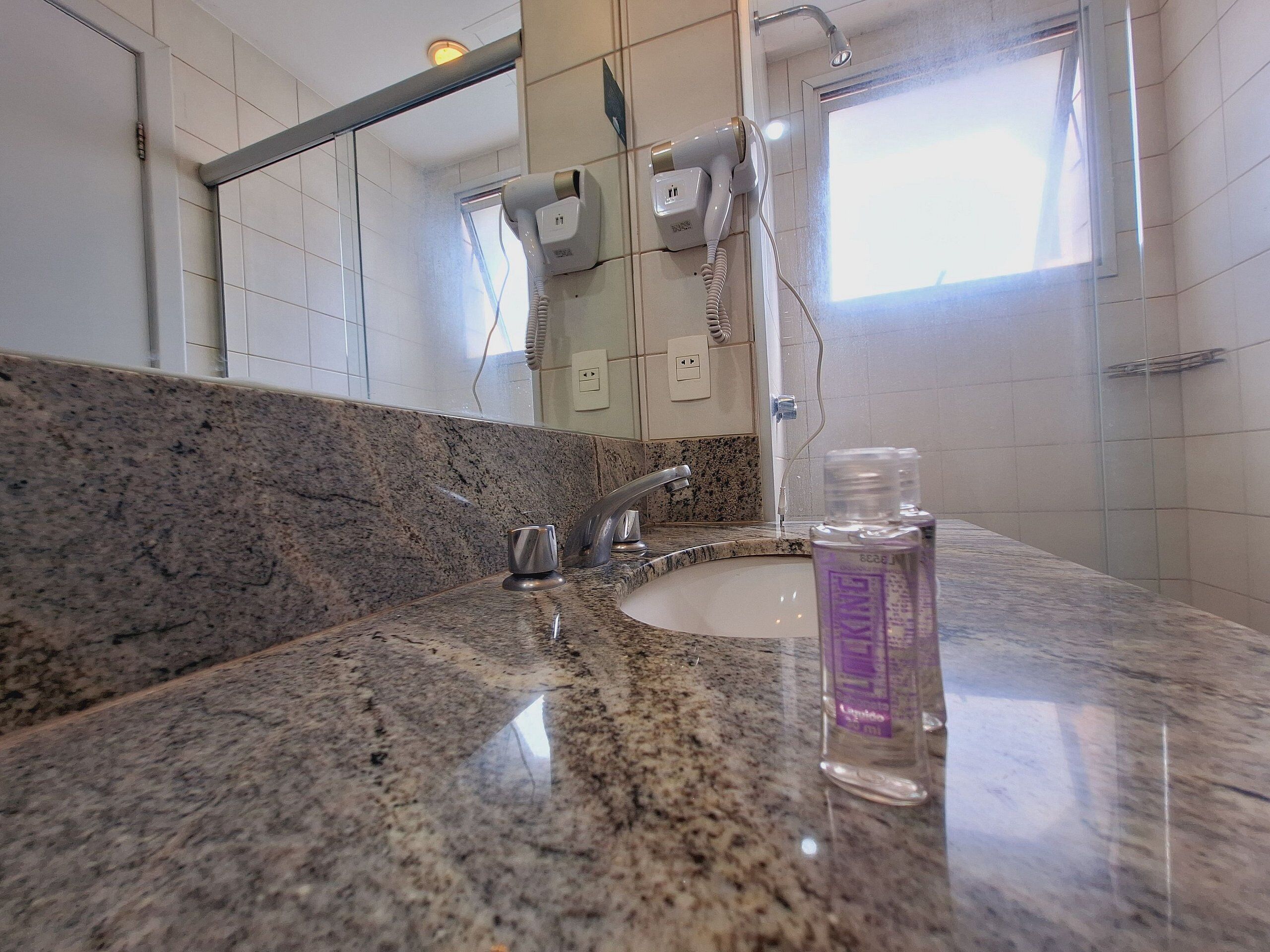 A bathroom with a granite countertop, a sink, a mirror, a handheld hair dryer, and a window.