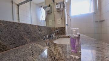 A bathroom with a granite countertop, a sink, a mirror, a handheld hair dryer, and a window.