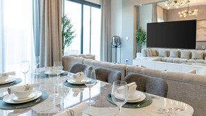 A modern living room with a dining area set for a meal, a large flat-screen TV, and a stylish chandelier. - Nasma Luxury Stays- Ras Al Khaimah (Ras Al Khaimah)