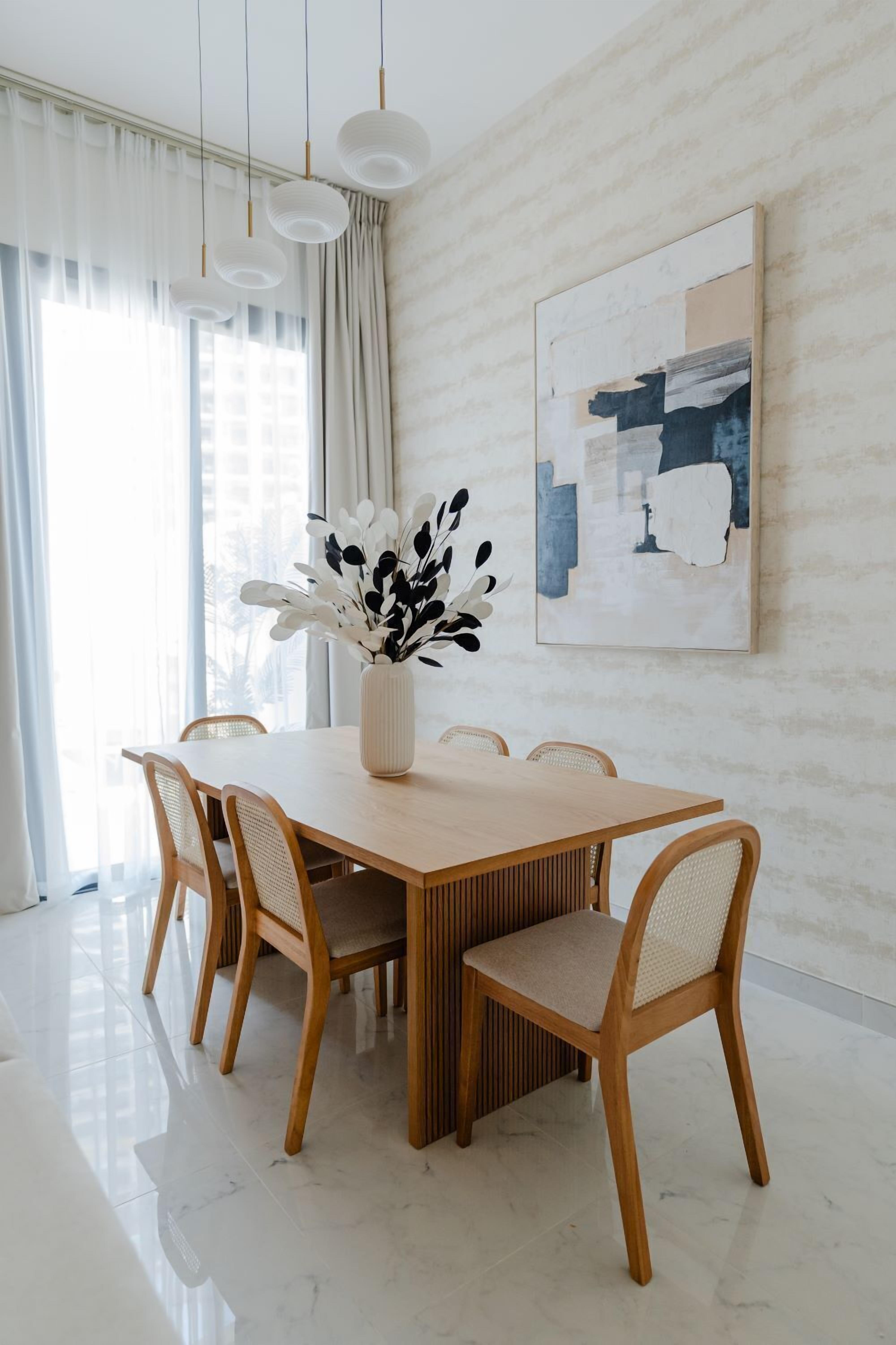 A dining area with a wooden table, four chairs, a vase with white flowers, and a modern abstract painting on the wall.