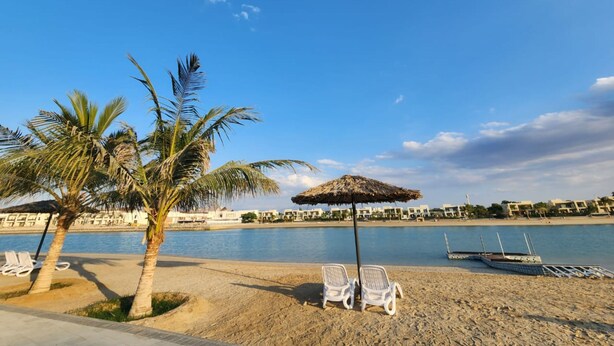 A beach with palm trees, a thatched umbrella, and lounge chairs. - Nasma Luxury Stays- Ras Al Khaimah (Ras Al Khaimah)