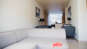 A modern living room with a light-colored sofa, a coffee table, and two floor lamps. There is a brick wall and a window in the background. - Spacious vacation home 5 min steps from city center. (Brugge)