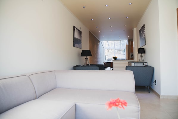 A modern living room with a light-colored sofa, a coffee table, and two floor lamps. There is a brick wall and a window in the background.