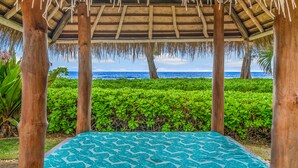 A covered outdoor seating area with a patterned blue cushion, wooden pillars, and a view of the ocean.