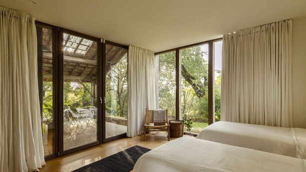 Un dormitorio con dos camas, piso de madera, un amplio ventanal corredera que da a un espacio exterior con una mesa y sillas, y vistas a árboles.