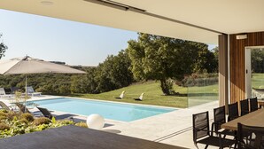 Un área de piscina exterior moderna con una mesa y sillas de comedor, rodeada de follaje y una vista despejada de un campo de golf.