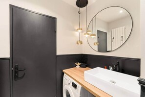 Une salle de bain moderne avec une porte sombre, un plan de travail en bois, un lavabo blanc, un lave-linge et un miroir rond agrémenté de luminaires suspendus.