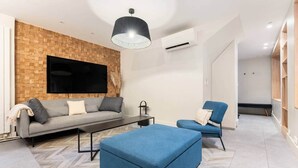 A modern living room with a grey sofa, blue armchair, and ottoman, a flat-screen TV mounted on a brick wall, and a pendant light. - Eiffel Tower - 4 bedrooms Air Conditioning (Paris)