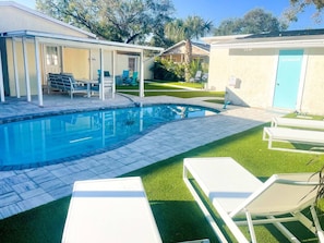 Una piscina con un patio techado, un baño y un césped con sillas de descanso.