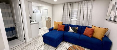 Una sala de estar moderna con un sofá seccional azul, una mesa de centro redonda y una cocina blanca al fondo.