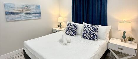 A neatly arranged bedroom with a large bed, two bedside tables, a blue curtain, and a painting on the wall.