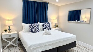 Een net opgemaakt bed met witte lakens en blauw gestreepte kussens, met aan weerszijden nachtkastjes met lampen en kleine planten.