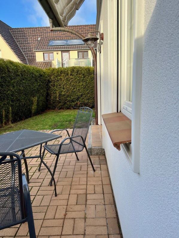 En terrasse med et bord og stole, en lille træhylle og en hvid væg.