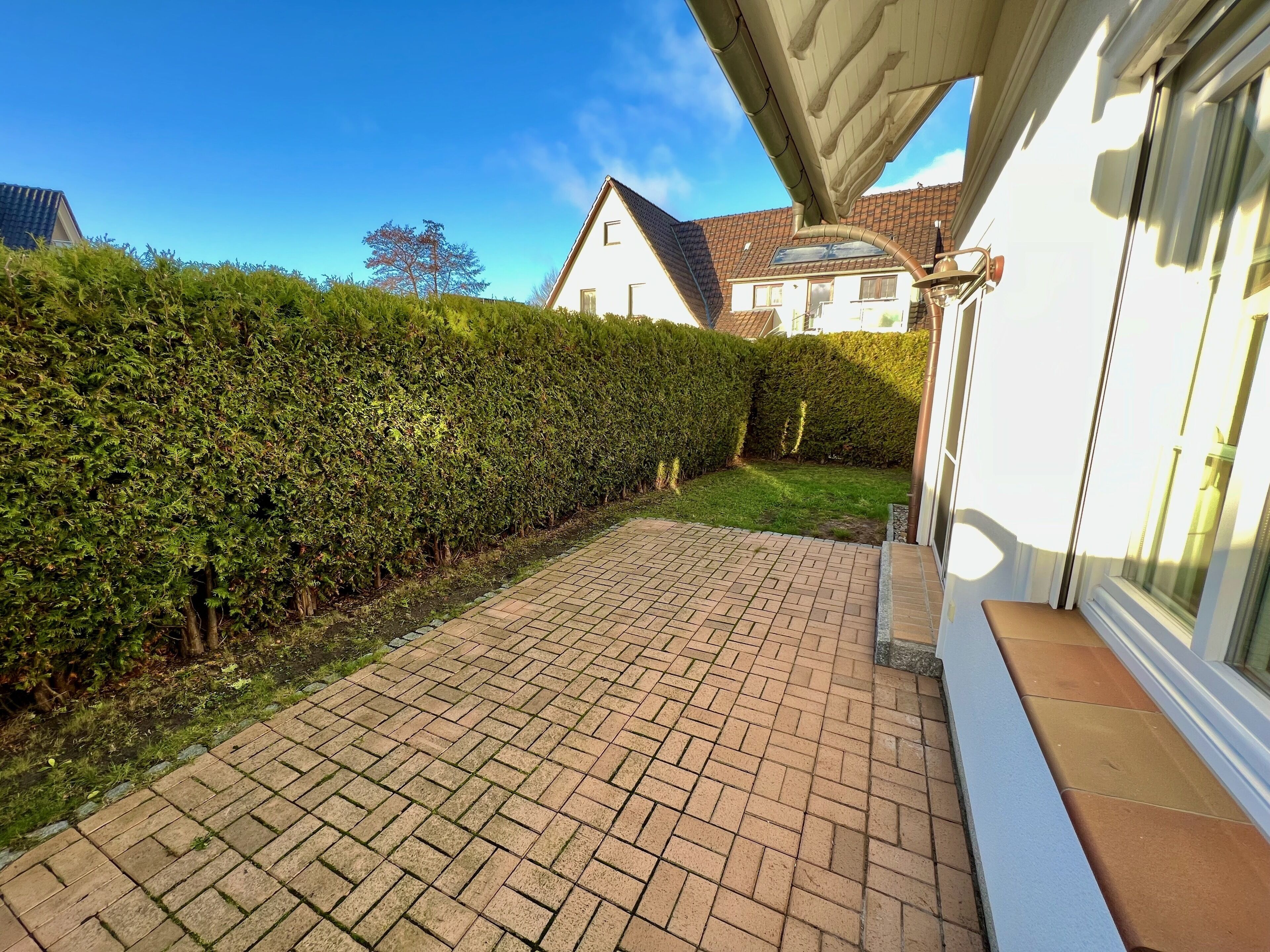 Une terrasse dallée, bordée d’une haie, avec une maison en arrière-plan.