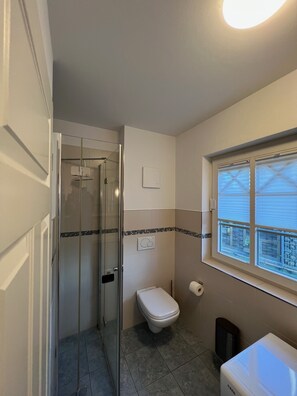 A bathroom with a glass-enclosed shower, a toilet, a window with blinds, and a white washing machine.