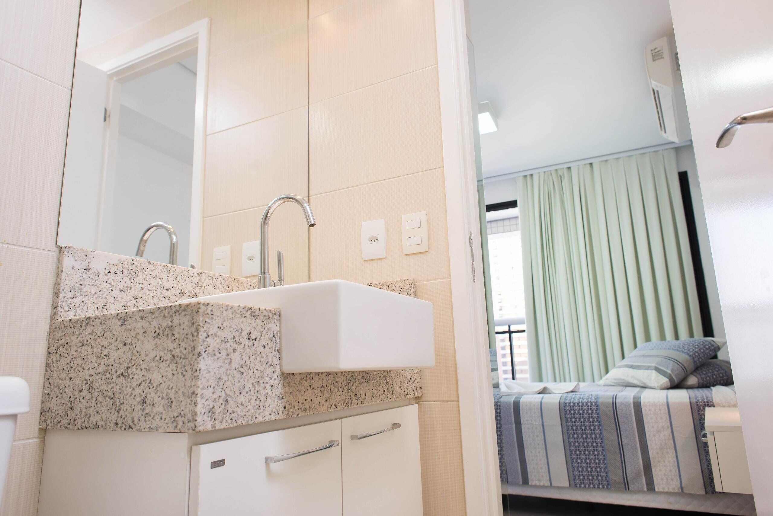 A modern bathroom with a granite countertop sink, a mirror, and a door leading to a bedroom with a bed and curtains.