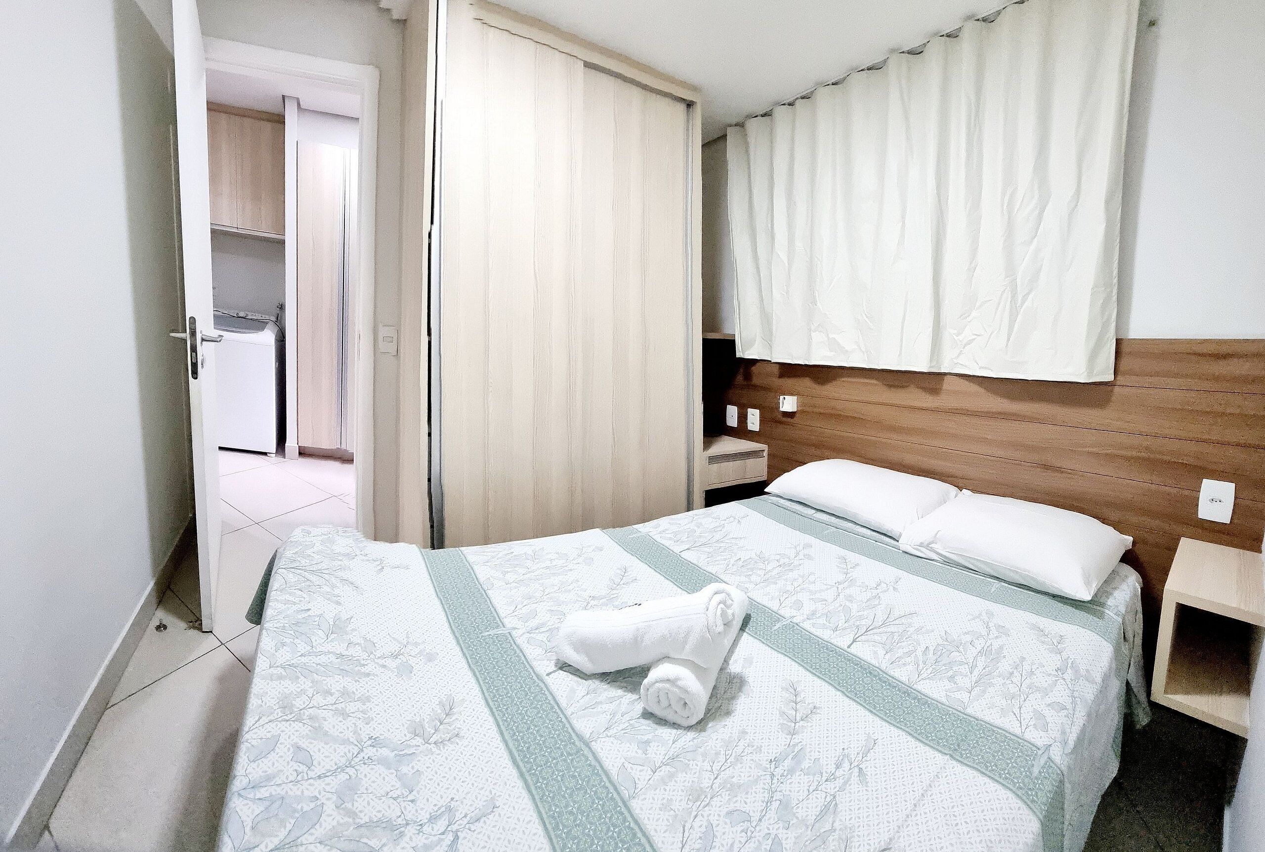 A hotel room with a bed, a wooden headboard, a nightstand, a white curtain, and a door leading to a bathroom.