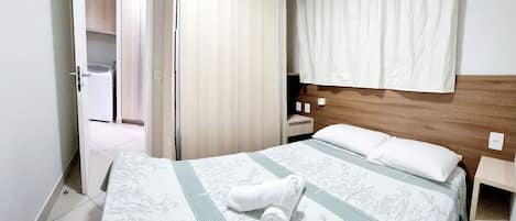 A hotel room with a bed, a wooden headboard, a nightstand, a white curtain, and a door leading to a bathroom.