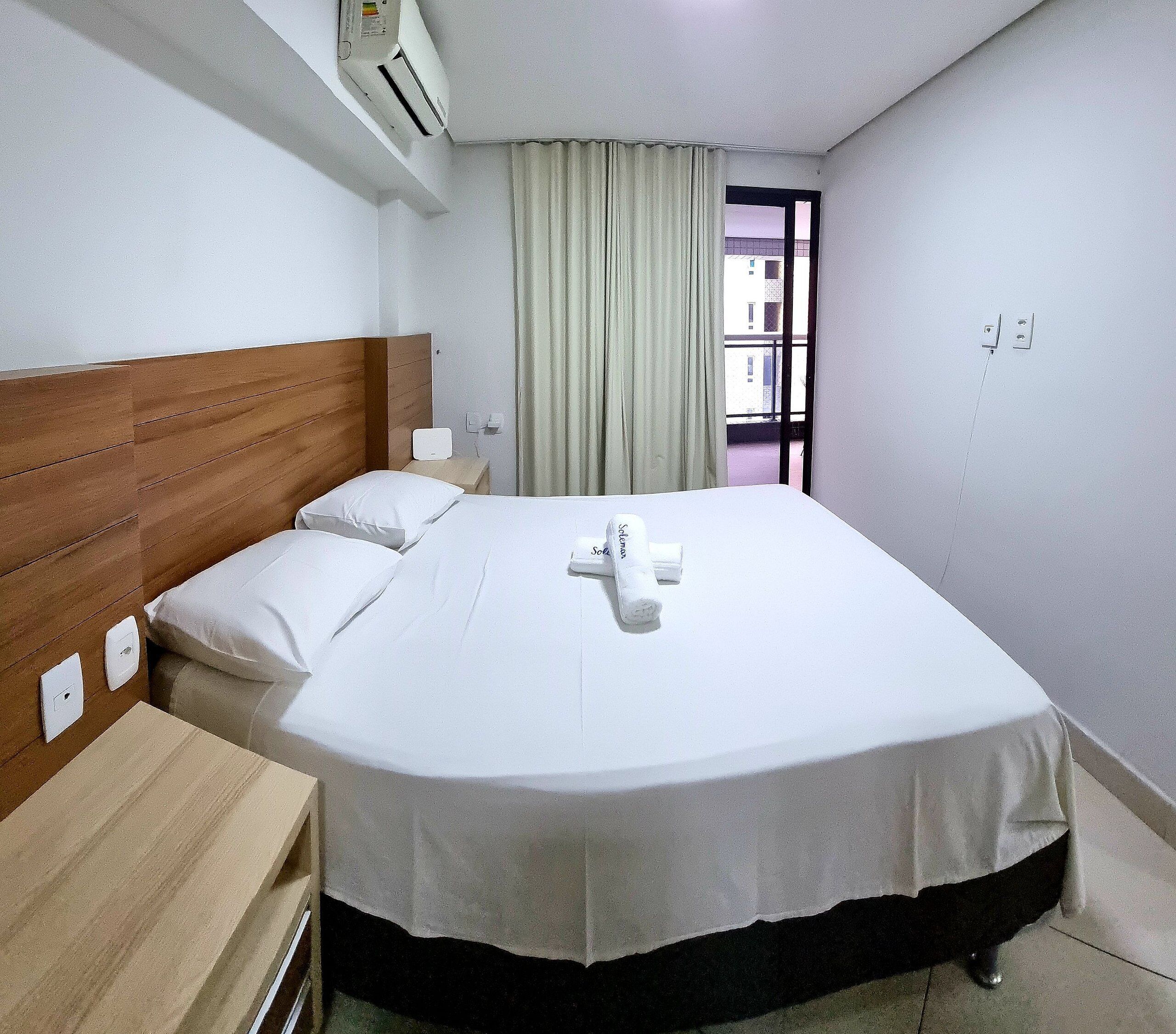 Une chambre d’hôtel avec une tête de lit en bois, un lit avec du linge de maison blanc, une table de chevet, une commode, un appareil de climatisation, une fenêtre avec des rideaux et une vue sur les bâtiments environnants.