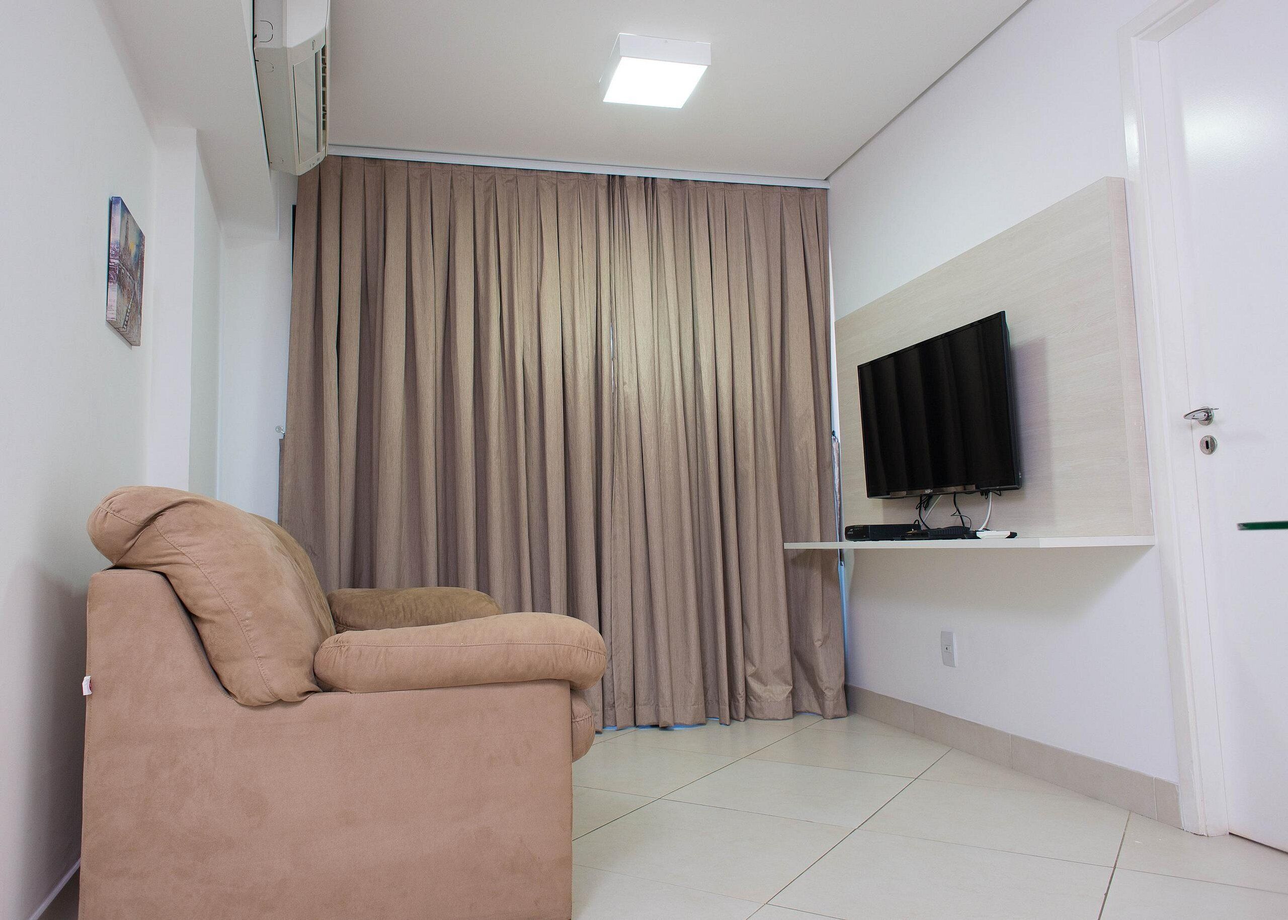 Una habitación con un sofá color beige, un televisor montado en la pared y un cortinero grande.