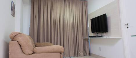 Una habitación con un sofá color beige, un televisor montado en la pared y un cortinero grande.