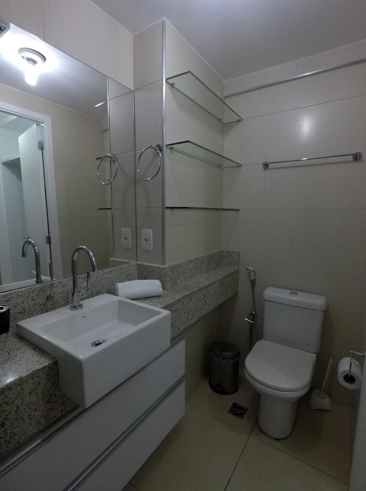 Casa de banho moderna com lavatório branco, espelho e sanita. Existem prateleiras de vidro acima do lavatório e um porta-toalhas.