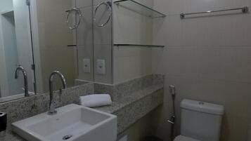 Casa de banho moderna com lavatório branco, espelho e sanita. Existem prateleiras de vidro acima do lavatório e um porta-toalhas.
