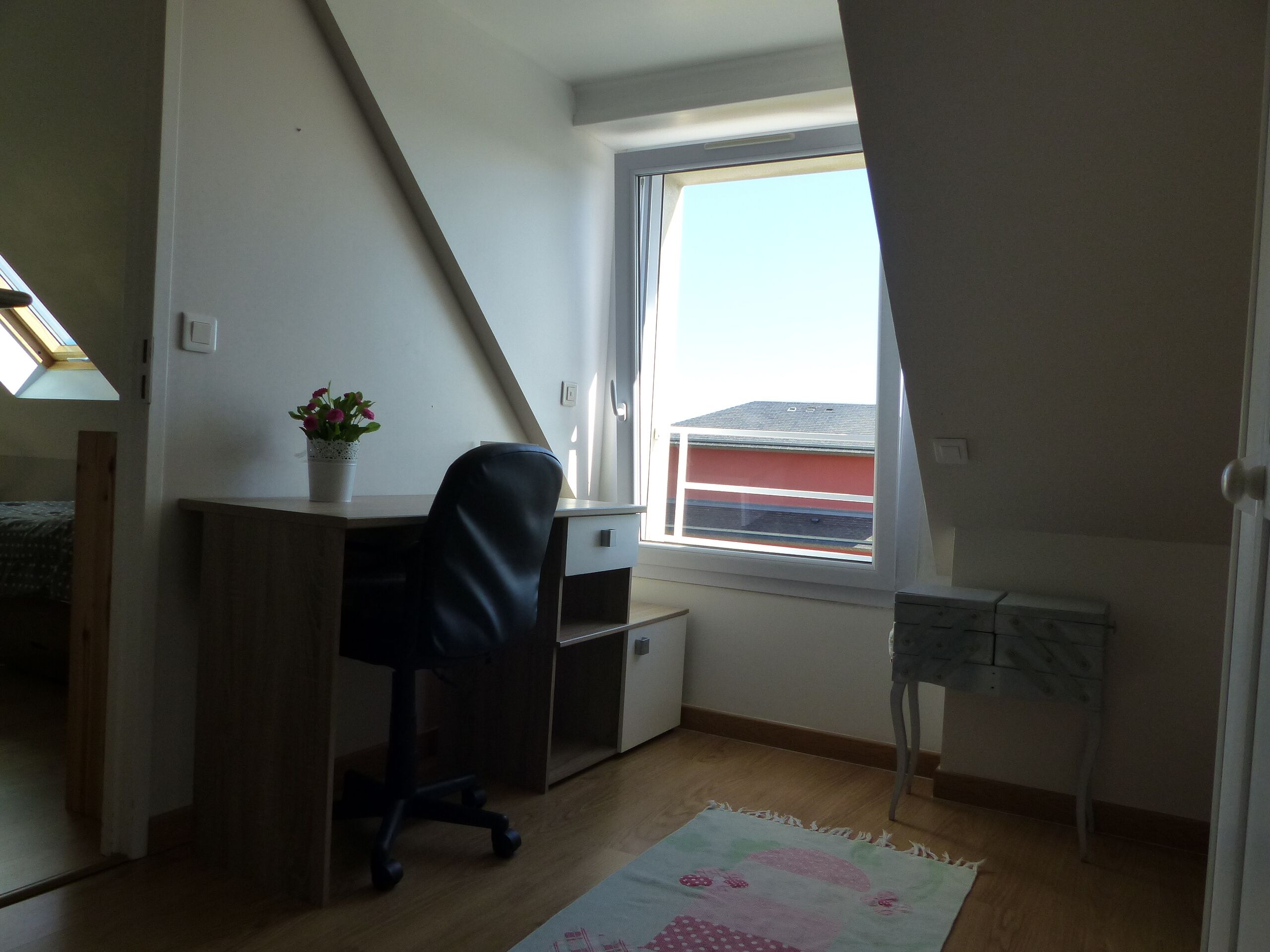Un pequeño y luminoso cuarto de ático con escritorio, silla y ventana con vistas a un edificio rojo.