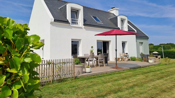 Een wit huis met een grijs dak, een houten terras en een rode parasol.