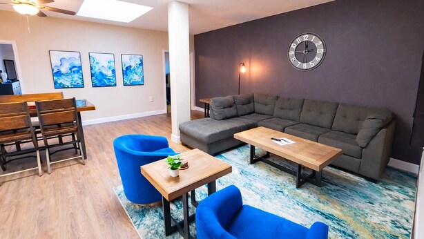 Ein modernes Wohnzimmer mit einer Eckcouch, einem Holztisch und blauen Sessel. Es gibt einen Essbereich mit Holztisch und -stühlen sowie einen Deckenventilator.