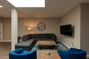 A living room with a sectional sofa, a coffee table, and two blue armchairs. There is a wall-mounted TV and a clock.