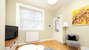 Un salon avec un téléviseur posé sur un meuble en bois, une petite table ronde avec deux tabourets, un fauteuil gris, une lampe sur pied et un tableau accroché au mur.