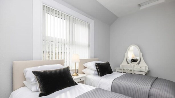 Une chambre d’hôtel avec deux lits, du linge de lit blanc, une table de chevet avec une lampe, un miroir et une fenêtre avec des stores.