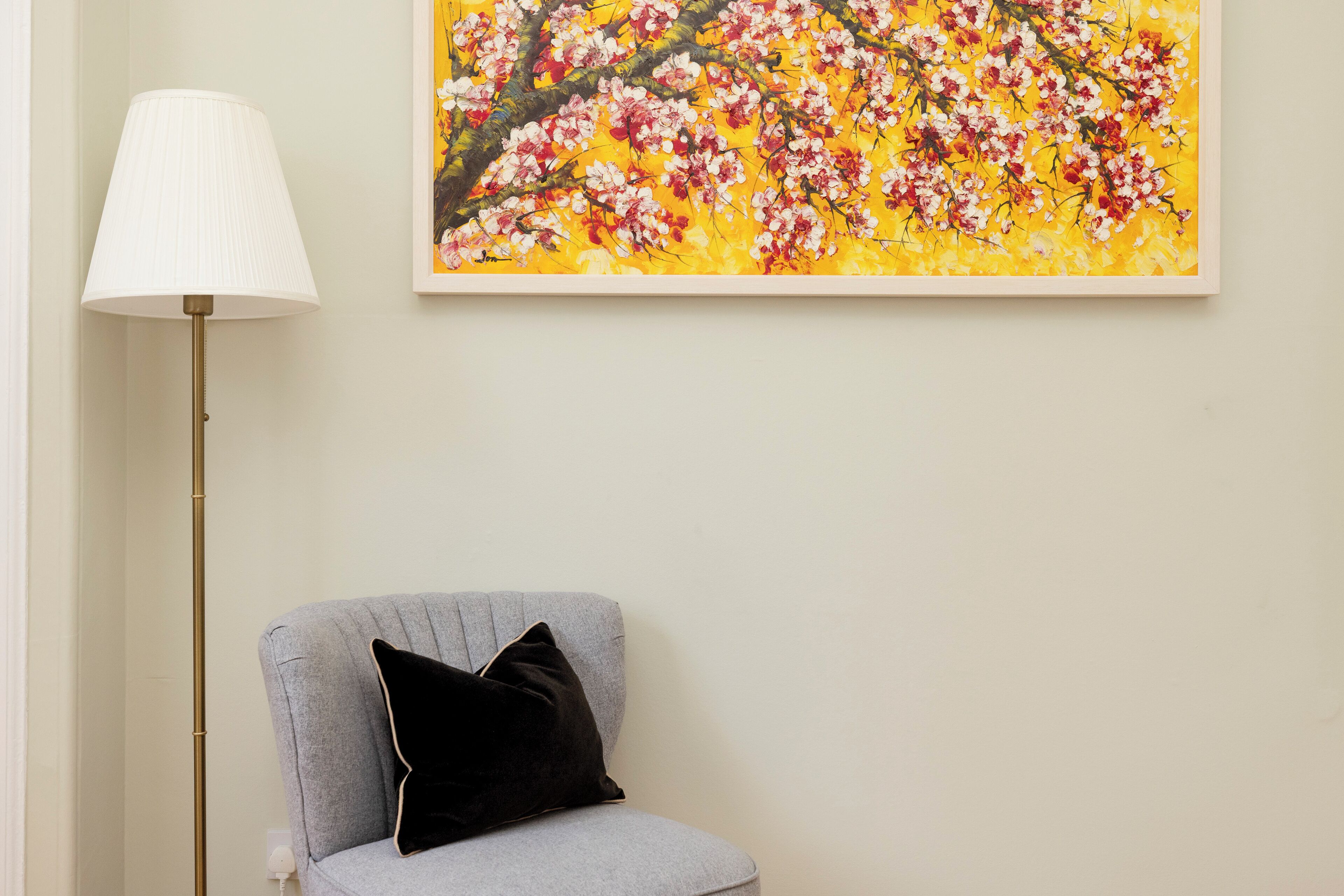 A room with a grey armchair, a black cushion, a floor lamp, and a framed painting of flowers on the wall.